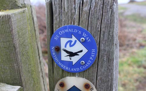 Circular sign on a wooden post highlighting St. Oswald’s Way – A Northumberland Coastal Path.