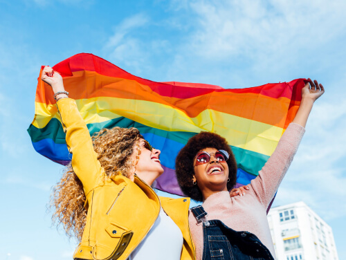 Two people smiling and holding a rainbow flag in celebration under a bright blue sky.