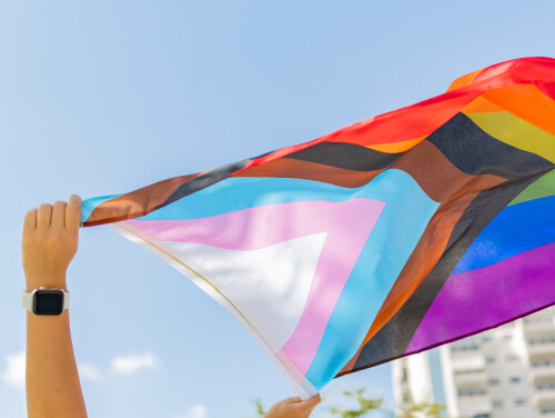 A hand holding a brightly coloured pride flag, waving in the breeze against a clear blue sky.