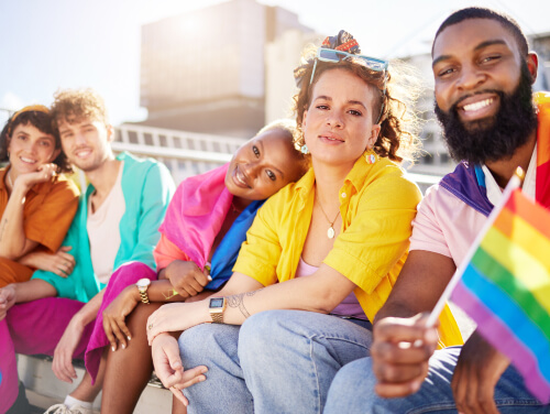 A group of five people smiling and sitting together, dressed in colourful clothing, with one person holding a rainbow flag.