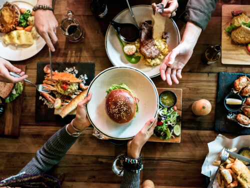 Overhead view of people sharing a meal with burgers, steak, seafood, and drinks on a wooden table.