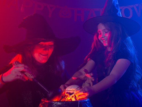 A mother and child dressed as witches pull spooky props from a pretend cauldron, garland reading Happy Halloween hangs on the wall behind them.