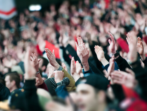 A crowd of people in the stands at a football game. They have their hands in the air ready to clap.