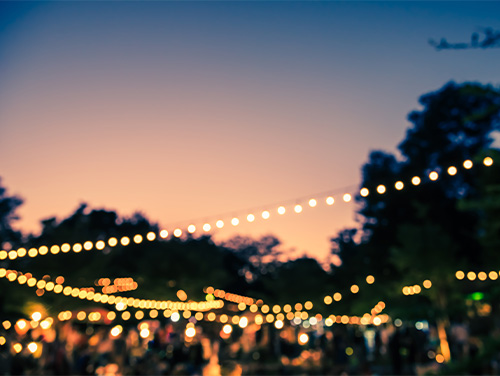 Dotted fairy lights under a twilight sky above a blurred festival ground full of people.