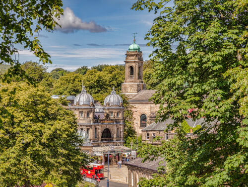 View of Buxton Opera House and its domed rooftops, framed by leafy green trees, with a historic clock tower and a red double-decker bus visible below.