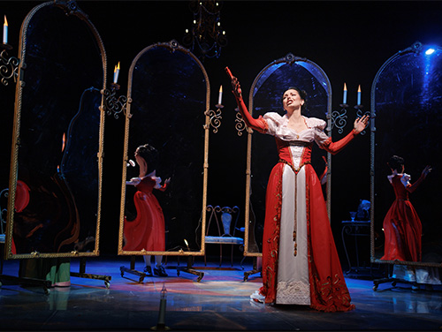 An actor playing a princess standing among several mirrors on a stage.