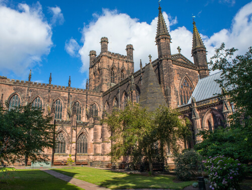 xterior view of Chester Cathedral, featuring intricate Gothic stonework, pointed arches, tall spires and historic towers, surrounded by landscaped gardens beneath a blue sky.