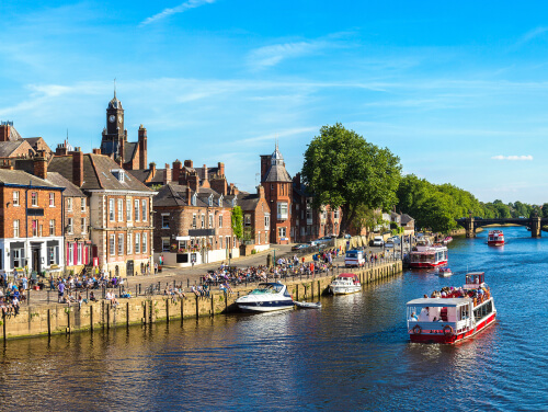 York riverside with historic buildings, boats on the River Ouse, and a sightseeing cruise passing through the city centre.