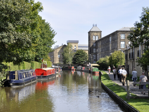 Skipton canal on a sunny day with river boats on one side and people on the other.