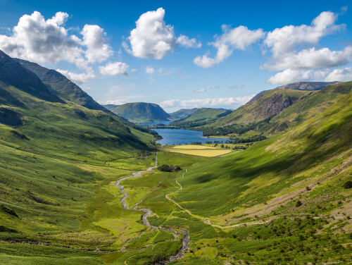 Green mountain valley with a winding stream leading to a lake under a blue, cloudy sky.