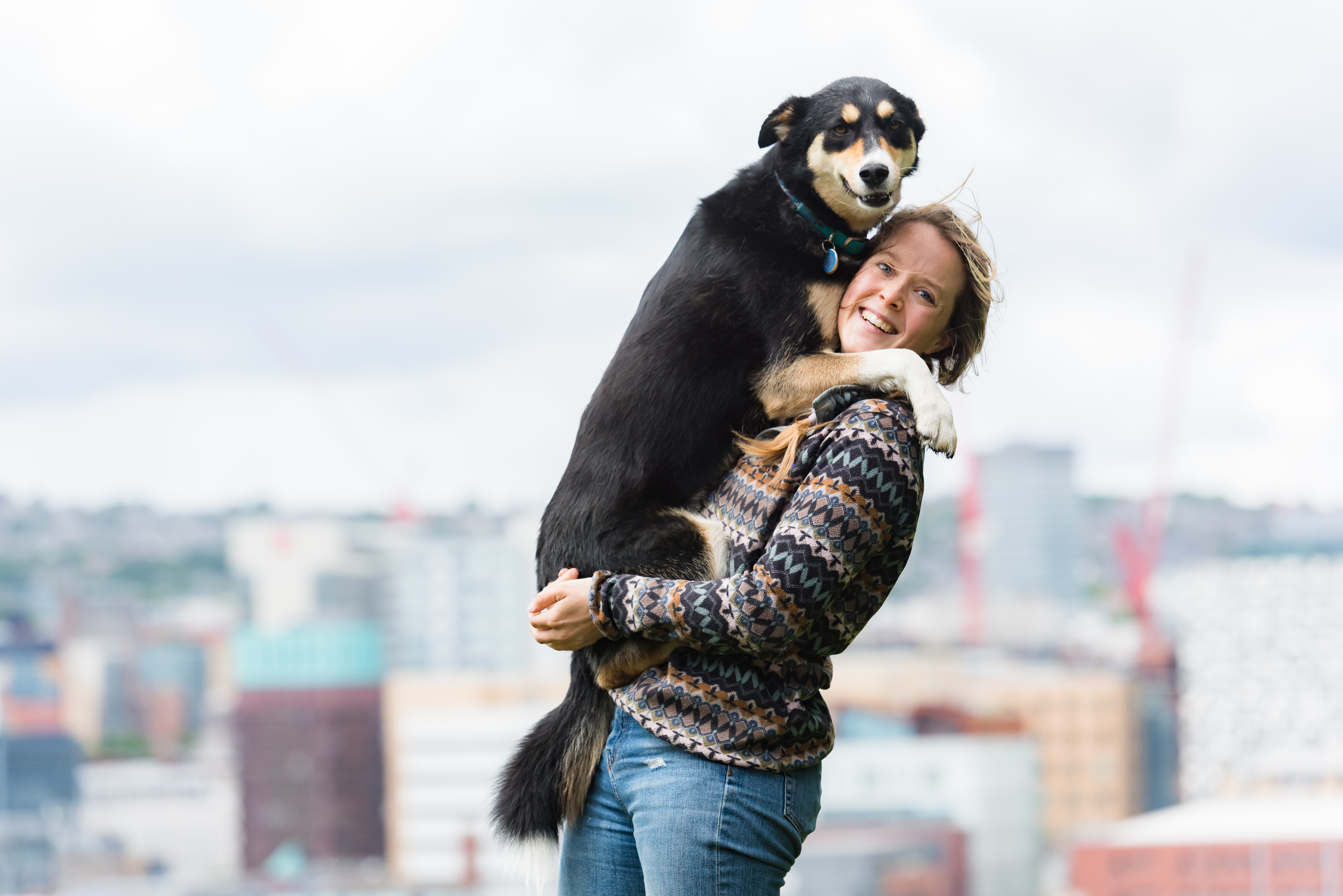 A woman lifts her dog up in a hug with the cityscape in the background