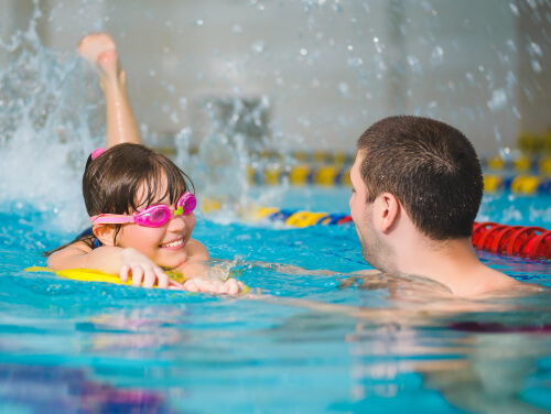 A girl swimming with her father in a pool and wearing bright pink goggles