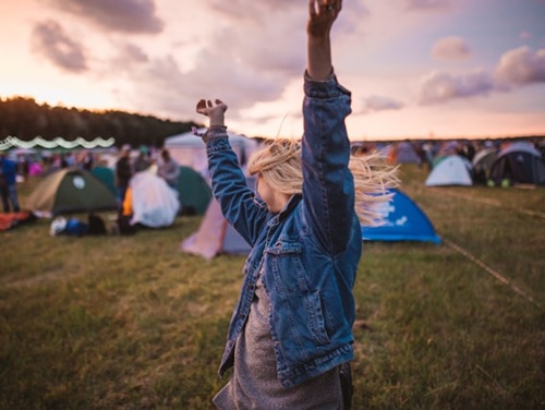 Blonde woman with their arms in the air standing in a field next to tents