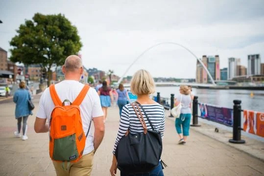People walking along Newcastle Quayside beside the River Tyne, with the Gateshead Millennium Bridge in the background.