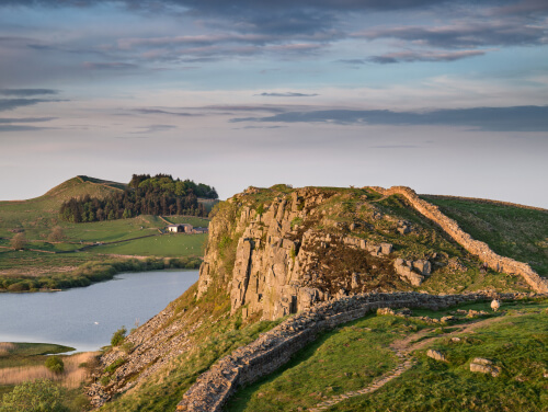 Hadrian's Wall disappearing over a blind summit, there is a body of water beneath the hill the wall sits on.