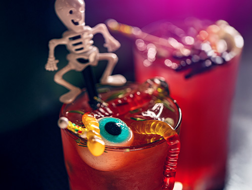 A red Halloween themed cocktail in a glass with a gummy candy eyeball, gummy worms, and a skeleton decorated straw.