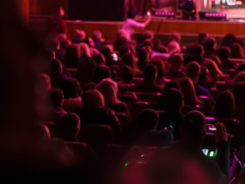 Audience seated in a theatre under red stage lighting.