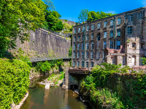 A historic building with broken windows next to a river, surrounded by greenery and a stone bridge.