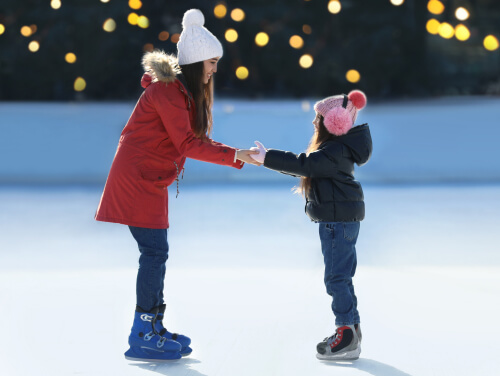 A teenager holding her little sisters hands as they stand in ice skates on a rink.