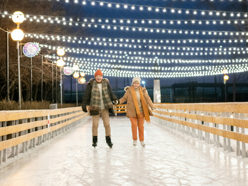 An older couple skating along a thin ice rink with fairy lights overhead.