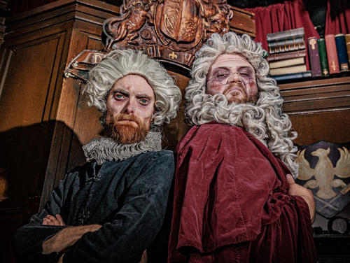 Two costumed performers in period wigs and robes pose in a wood-panelled room.