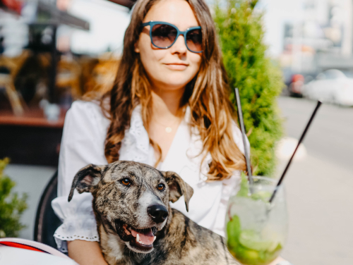 Woman sitting with a dog and a drink at an outdoor table in a city setting.