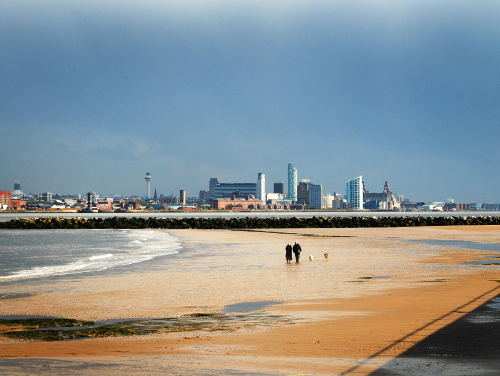 People walking a dog along New Brighton Beach, with the Liverpool skyline across the River Mersey.