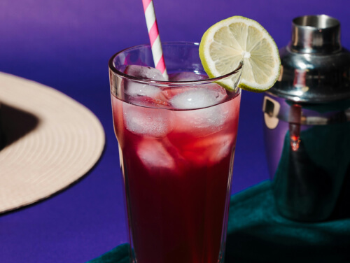 A Vimto cocktail with ice in a glass, garnished with a lime, sitting in front of a cocktail shaker.