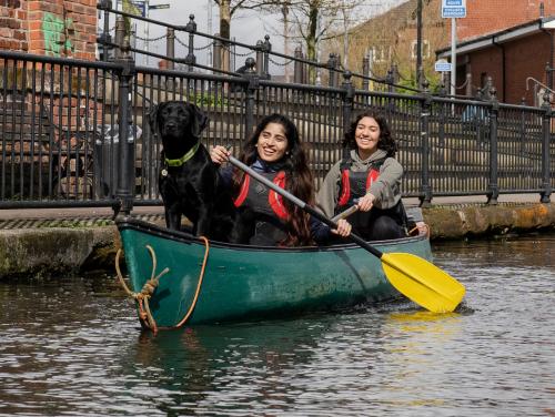 Two people canoeing along a Manchester canal with a black dog sitting at the front of the canoe.