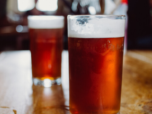 Two glasses of foamy ale sitting atop a wooden bar table.