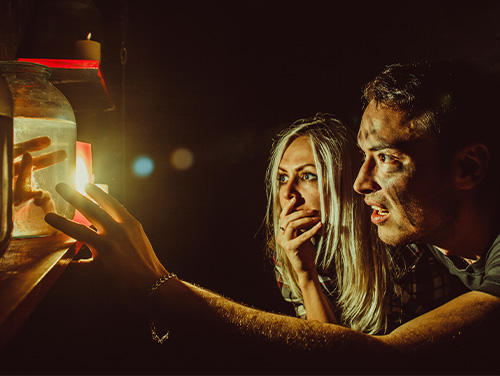 A couple in a dark room gaze in slight horror and intrigue at a fake hand in a jar. 