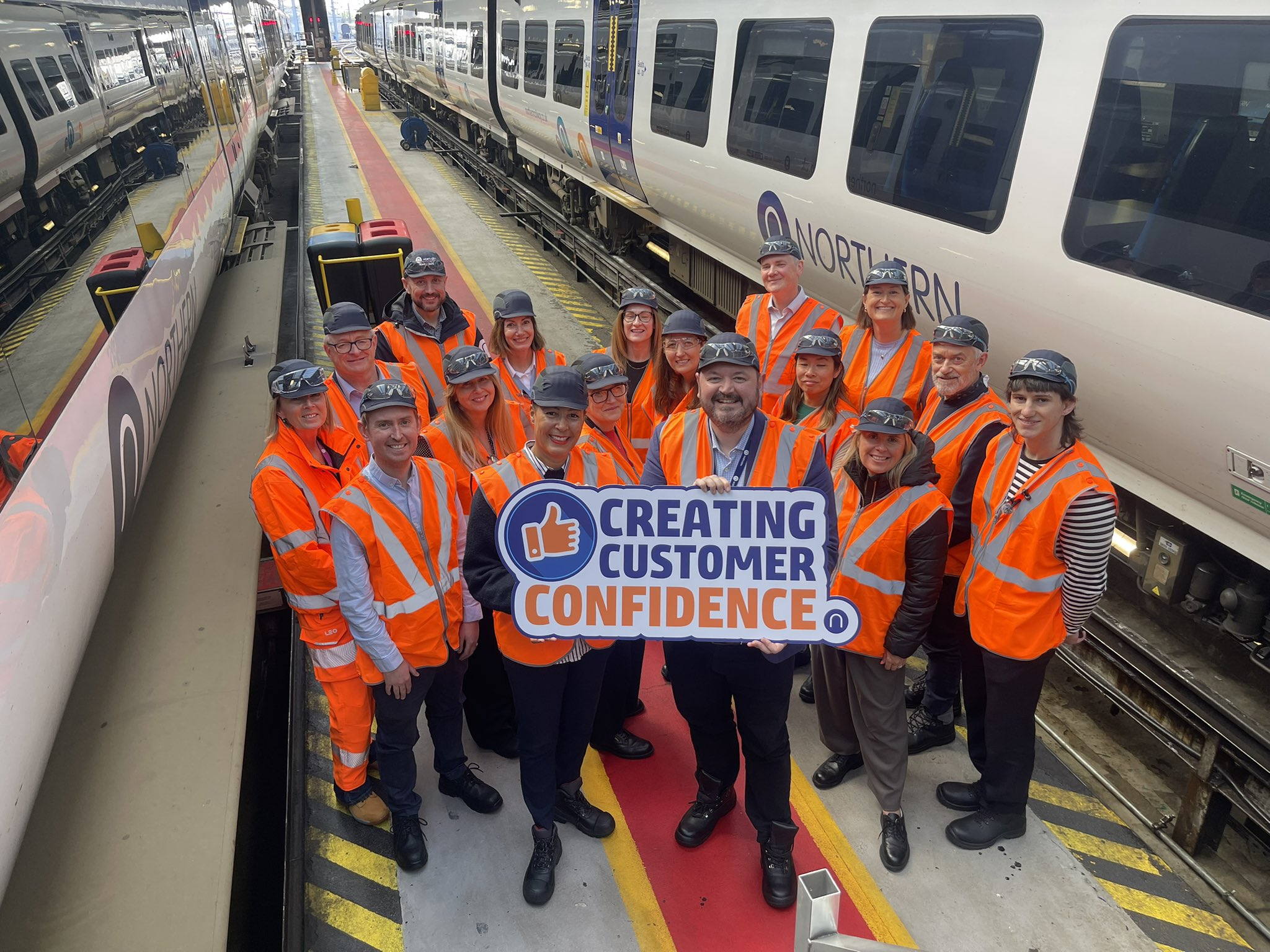 Photo shows a group of adults standing between two trains in a train care centre. Over their clothes they wear orange high vis vests and are wearing black railway caps. The front two of the group are holding up a sign which reads 'Creating Customer Confidence'