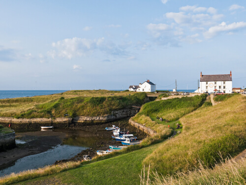 A coastal scene in Northumberland with boats docked at a harbour and a white house on a grassy hill.