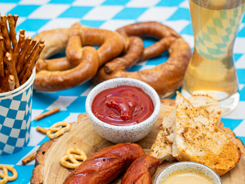 A selection of Oktoberfest themeds foods on a blue and white checkered table with Bratwurst sausages, pretzels, and beer.