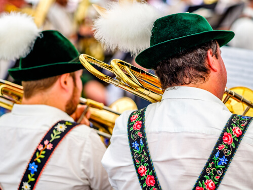 Two men facing away from the camera, wearing lederhosen, Bavarian hats, and blowing gold trumpets.