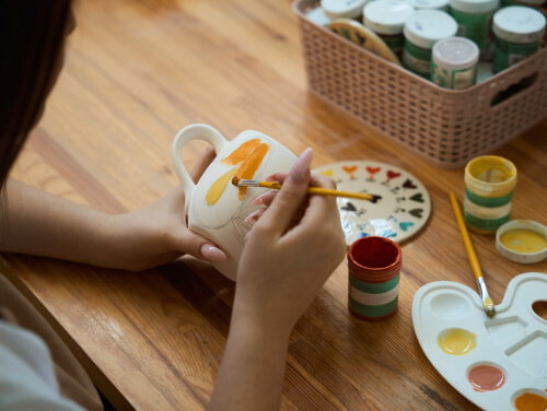 Person painting orange and yellow brushstrokes onto a ceramic mug at a craft table.