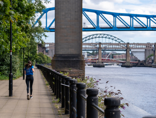 A runner following the riverside path beside the River Tyne in Newcastle, with several bridges visible in the background.
