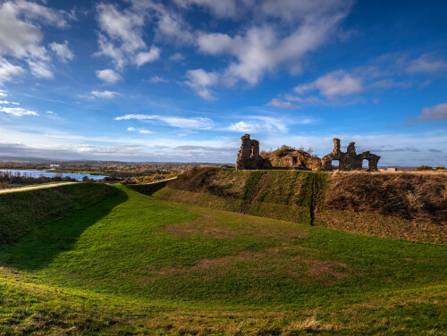 Ruins of a stone castle on a grassy hill under a blue sky, with countryside and water visible in the distance.