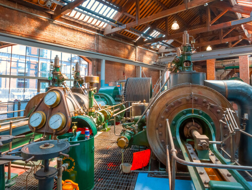 Historic industrial machinery with large metal turbines, pipes and gauges inside a brick building.