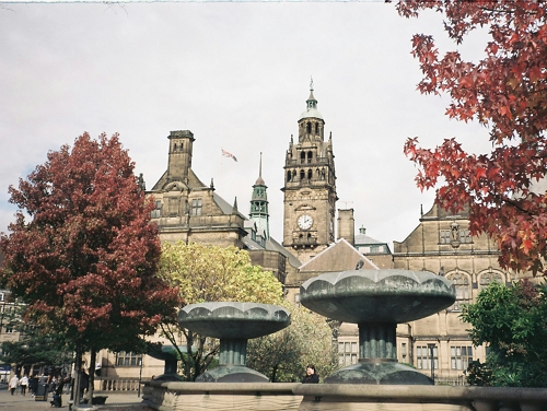 Historic stone building with a clock tower, framed by autumn trees and large fountains in the foreground.