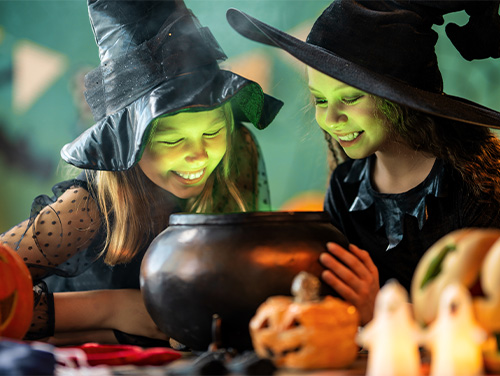 Two girls dressed up as witches for Halloween gaze into a plastic black cauldron, the green light from within the cauldron illuminating both their faces.