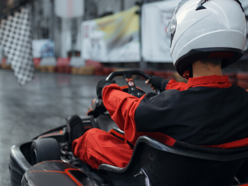 Person wearing a red and black racing suit driving a Go Kart on an indoor racing track.