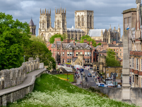 York city walls with York Minster in the distance, overlooking a road, greenery and spring flowers.