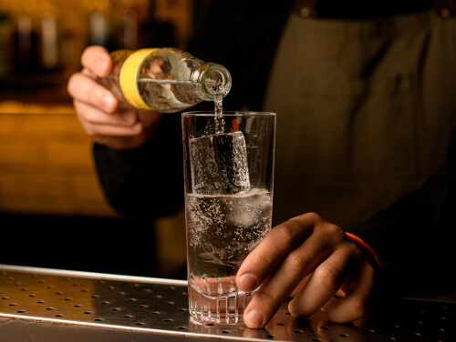 A bartender pouring tonic into a tall glass with ice at a bar.
