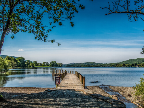 The image depicts a wooden pier extending into a calm lake, surrounded by lush greenery and under a clear blue sky.