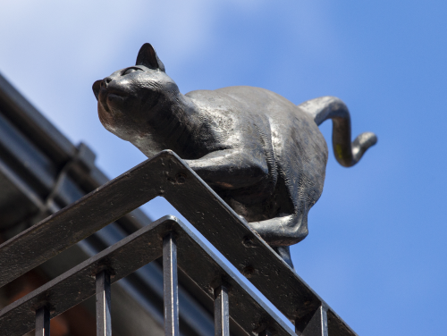 Bronze cat statue perched on a railing against a clear blue sky.