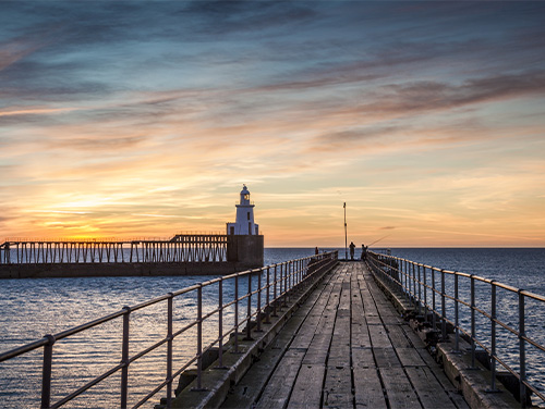 Blyth pier during sunrise. Two people sit at the end of the pier fishing.