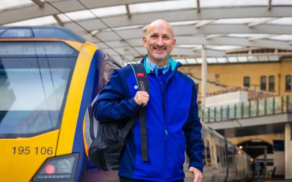 A photo of a male Northern train driver smiling on the platform in front of a 195 train. He is carrying a backpack over his right arm.