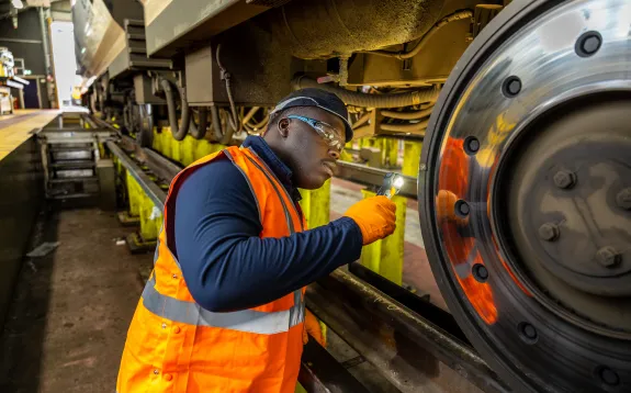 Image shows a male engineerworking on a train. He is wearing a long sleeved dark blue top with an orange hi-vis vest over the top, orange gloves, a blue cap and safety glasses. He is shining a torch at a train wheel. The train is lifted above him in the depot.