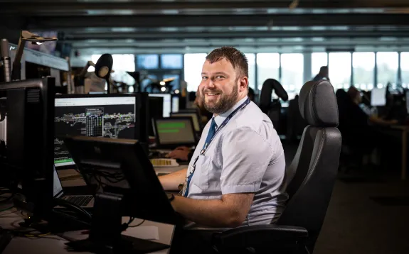 Image shows a man sitting in a control room in a black leather chair with his desk in front of him. There a three screens on his desk and he is looking to his left and smiling at the camera. He is wearing a pale blue short sleeved shirt, tie and a lanyard